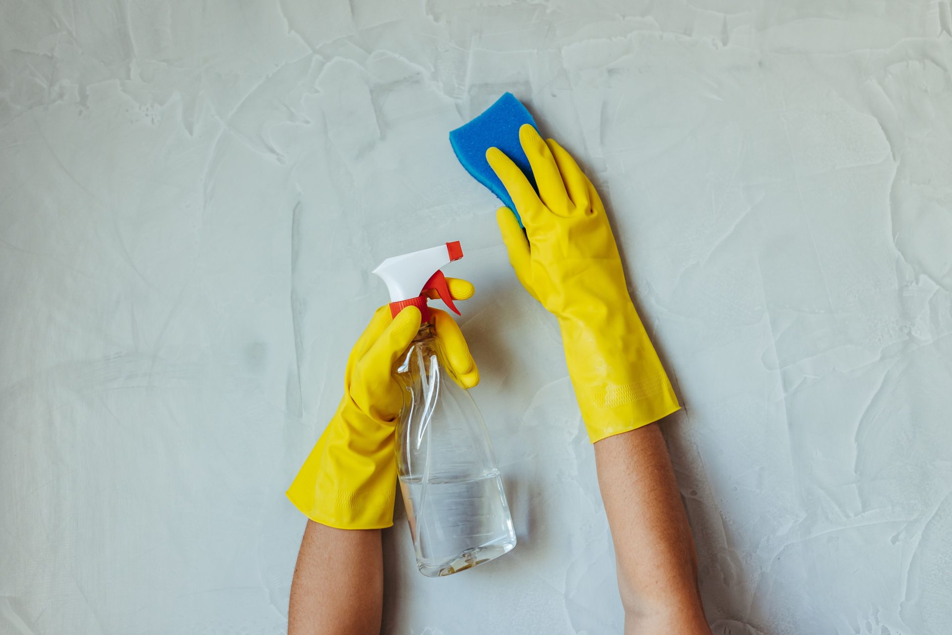 Nahaufnahme von Händen in gelben Handschuhen, die eine Wand mit einer Sprühflasche und einem Schwamm reinigen.